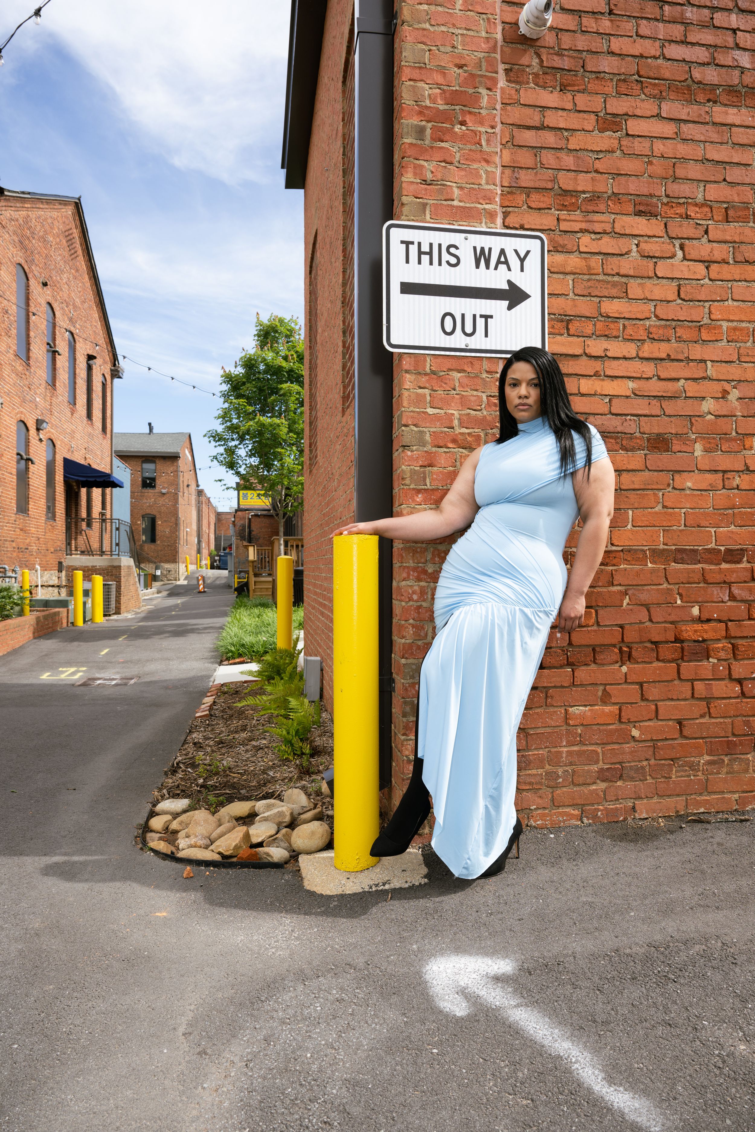 Pregnant woman in light blue dress posing by brick wall near This Way Out sign in urban alley.