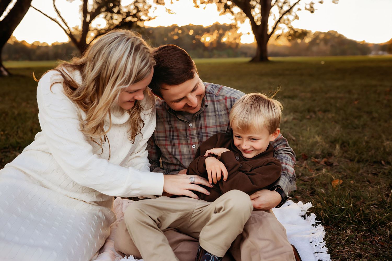 A joyful family of three enjoys a cozy outdoor picnic on a white blanket, with parents tickling their laughing young child.