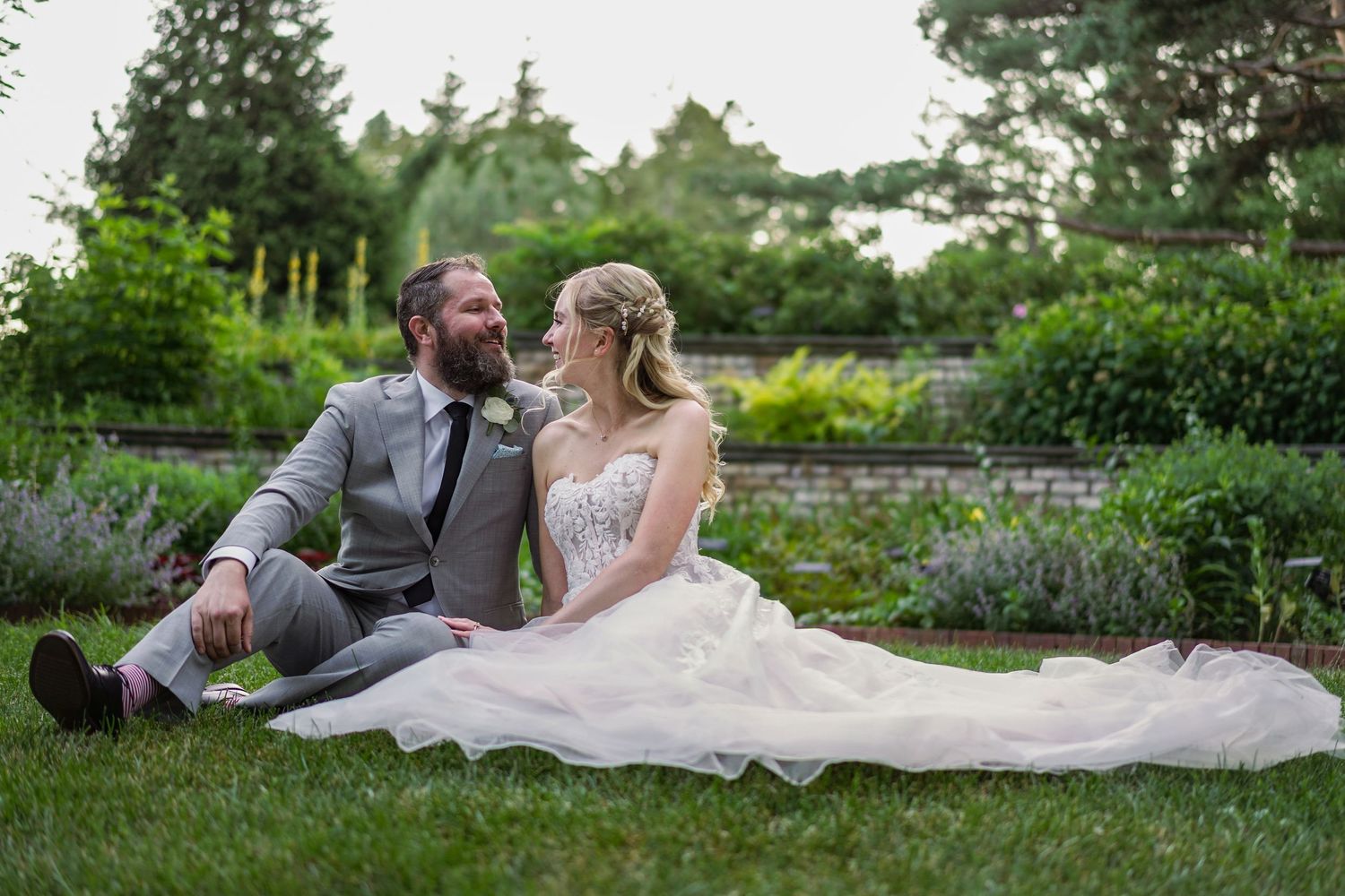 Bride and groom sitting on grass in a lush garden, gazing lovingly at each other.