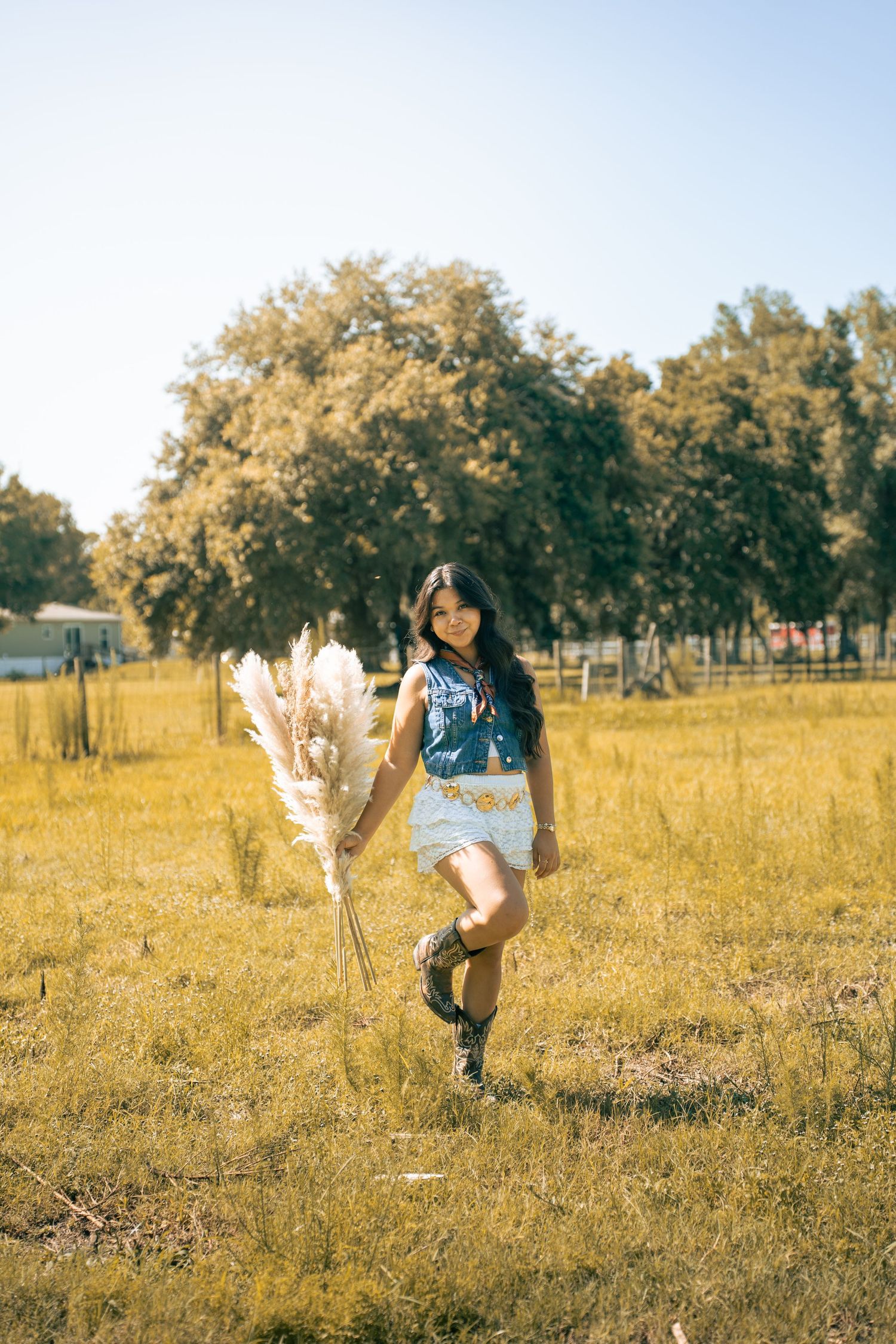 Young woman in denim vest, lace shorts, and cowboy boots holding pampas grass in a sunny field.