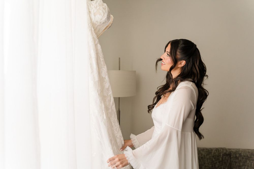 Bride in white robe admiring her lace wedding dress hanging by a bright window before the ceremony.