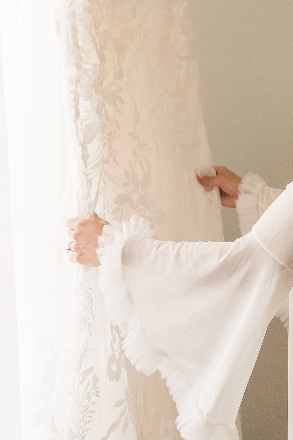 Bride in white robe holding floral lace wedding dress near a bright window on wedding morning.