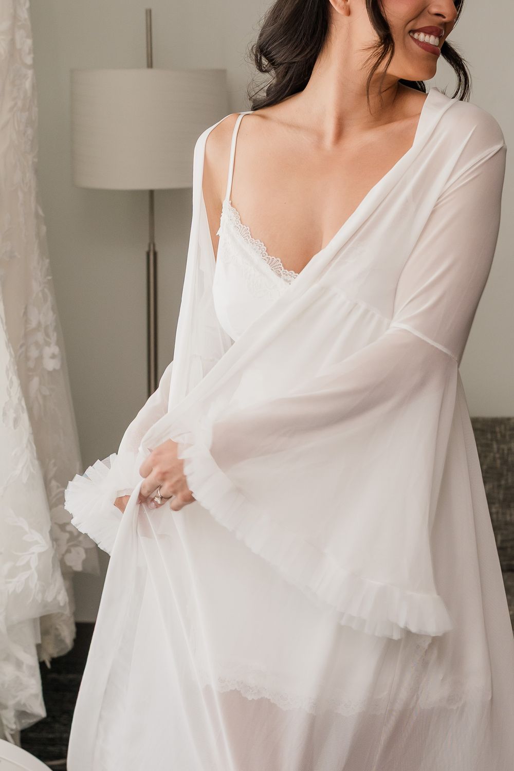 Smiling bride in white lace slip dress and sheer robe getting ready in a bright bridal suite.