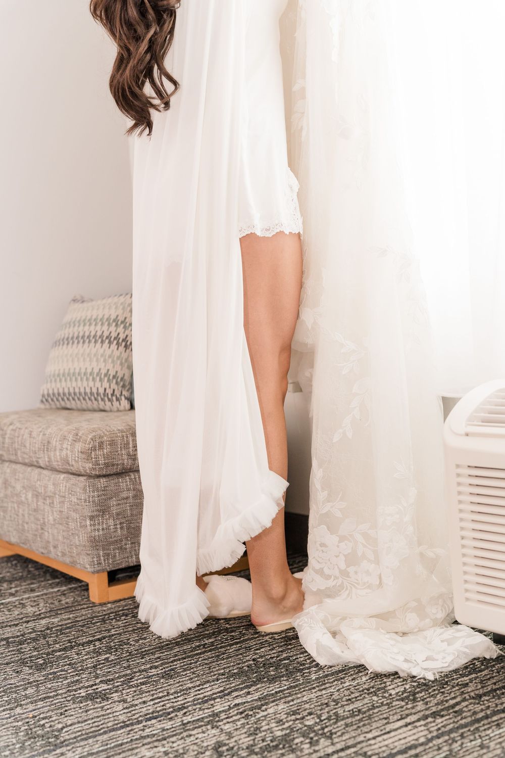 Bride in white robe standing beside lace wedding dress in bright hotel room on wedding day.