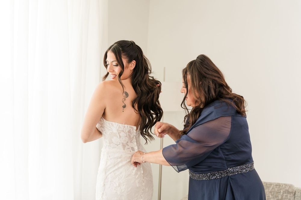 A woman in a lace wedding gown gets her dress fastened by another woman in a blue dress near a bright window.