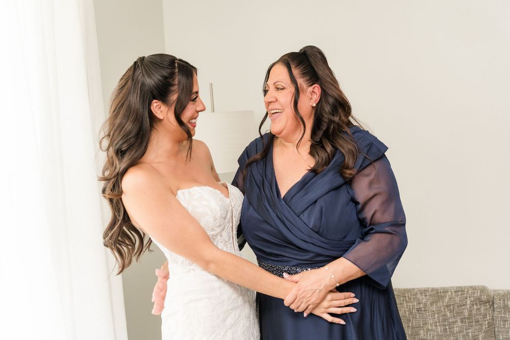 Bride in white lace gown sharing a joyful moment with her mother in navy blue dress before wedding.