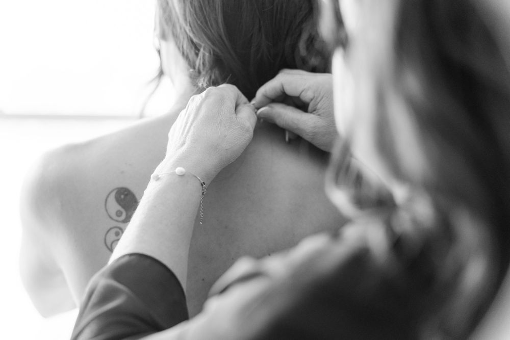 Black and white photo of a woman fastening a necklace on a bride with a tattoo on her back.