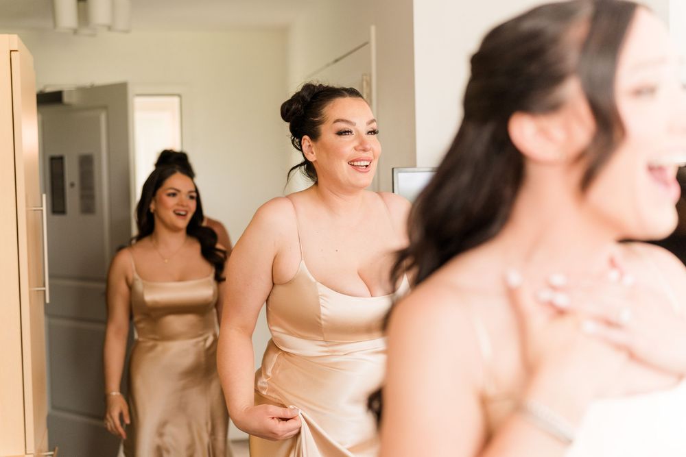 Bridesmaids laughing and smiling in gold satin dresses while helping bride get ready on wedding day.