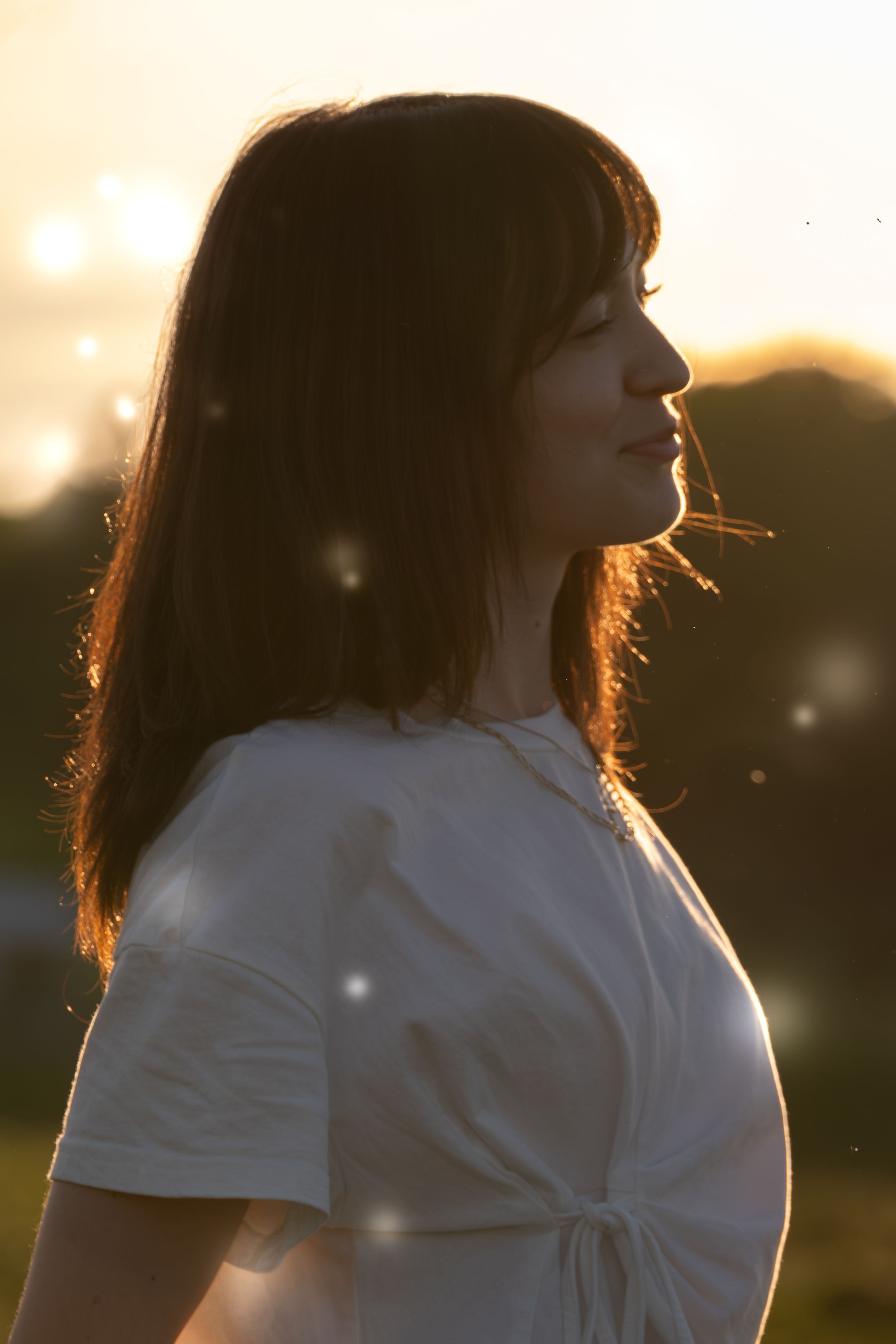 Woman in white t-shirt standing outdoors at golden sunset with soft bokeh light effects.