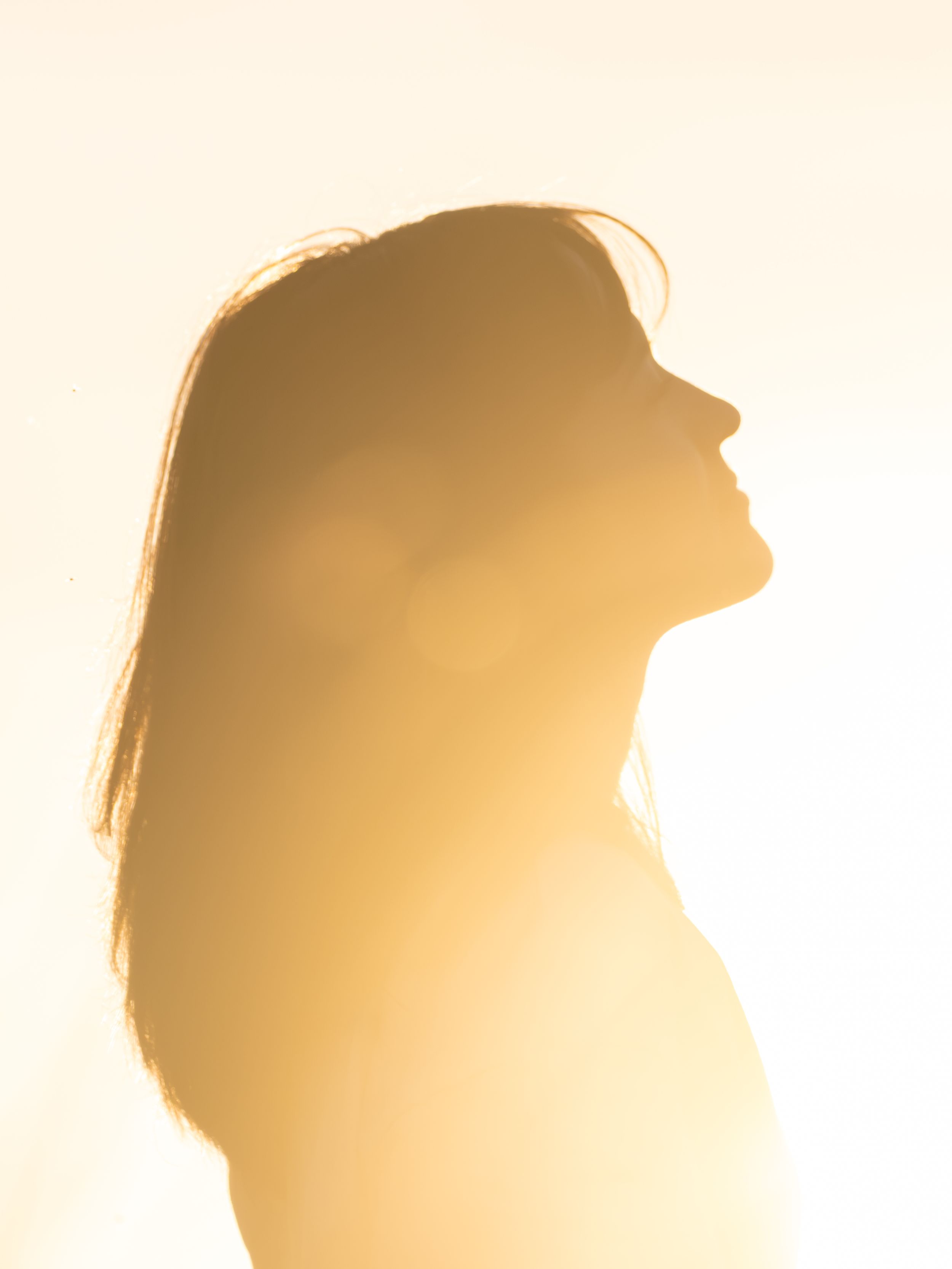 Golden backlit silhouette of a woman with long hair gazing upward against a bright warm glowing background.