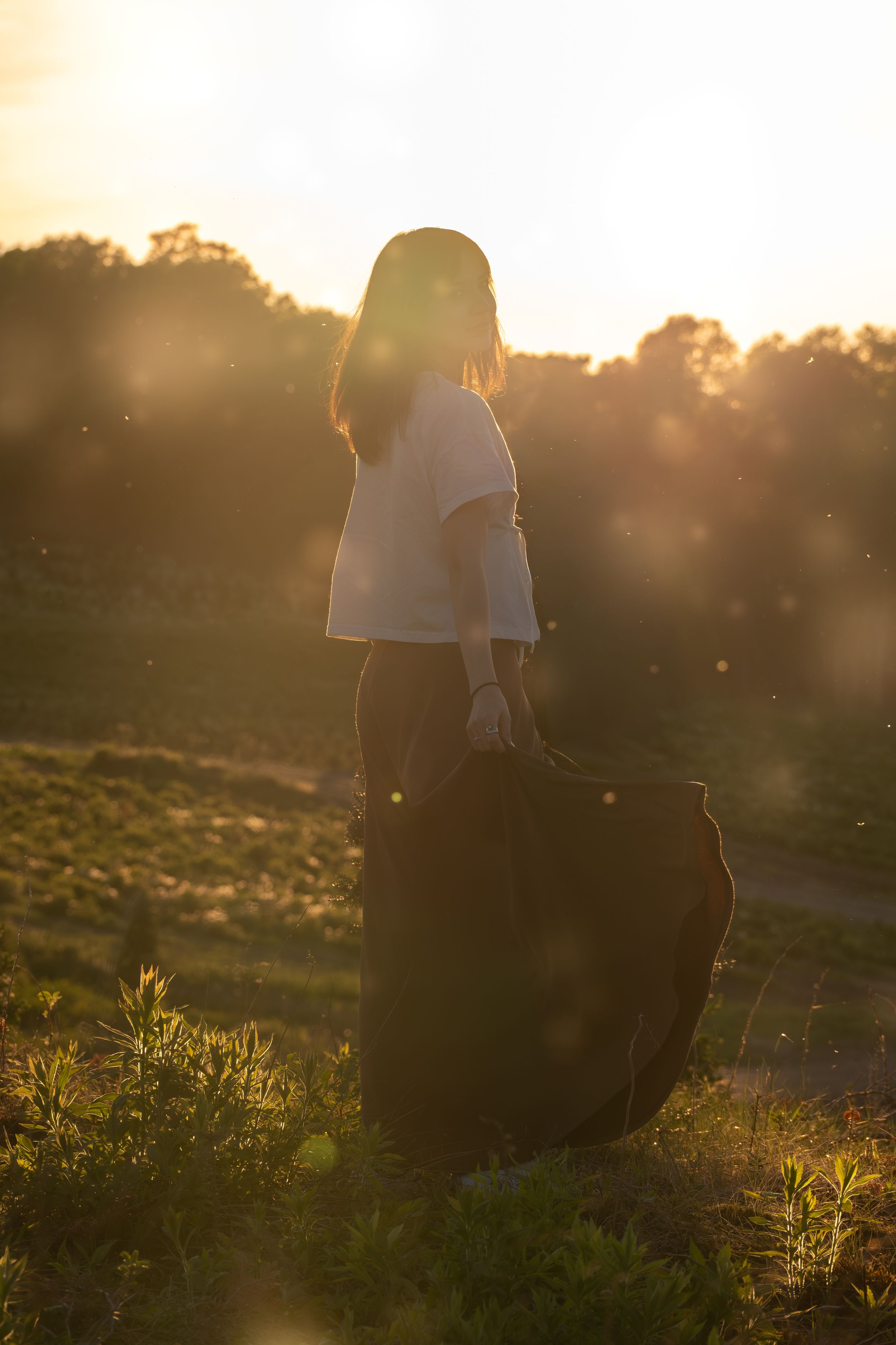 Woman in white top and black skirt standing in golden sunset light in a field with lens flare.