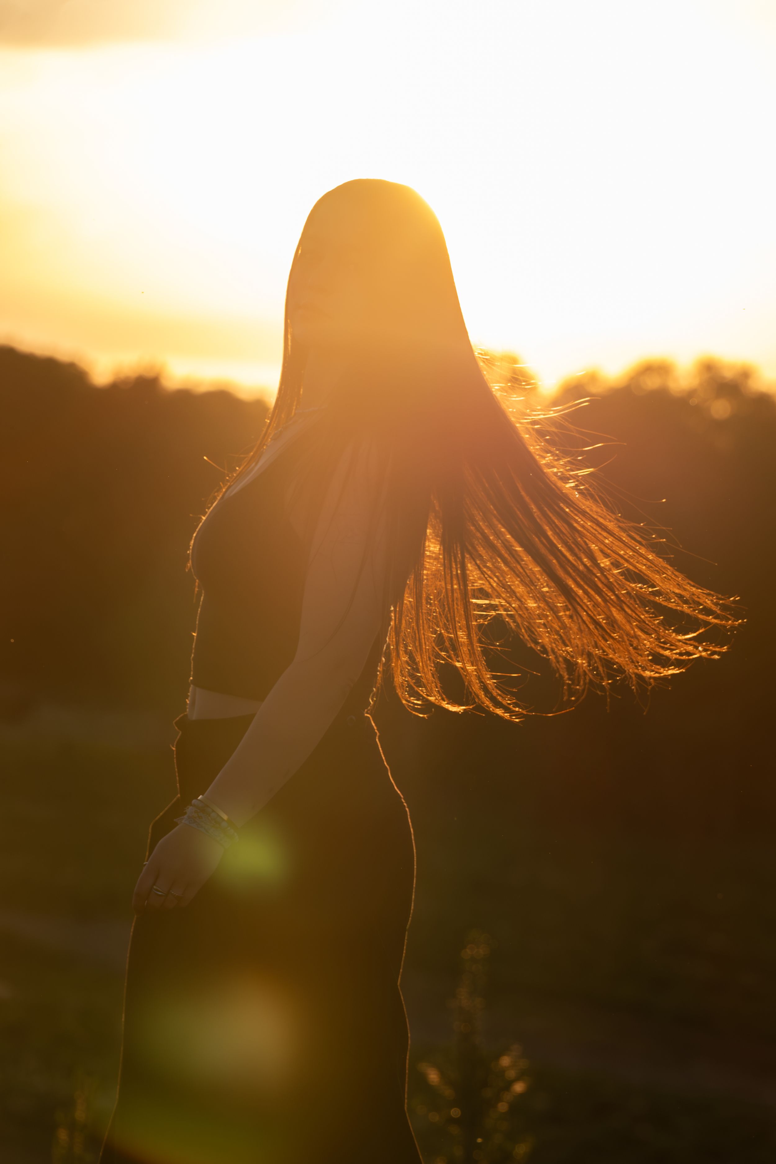 Silhouette of a woman with long flowing hair against a golden sunset backdrop in an outdoor field.