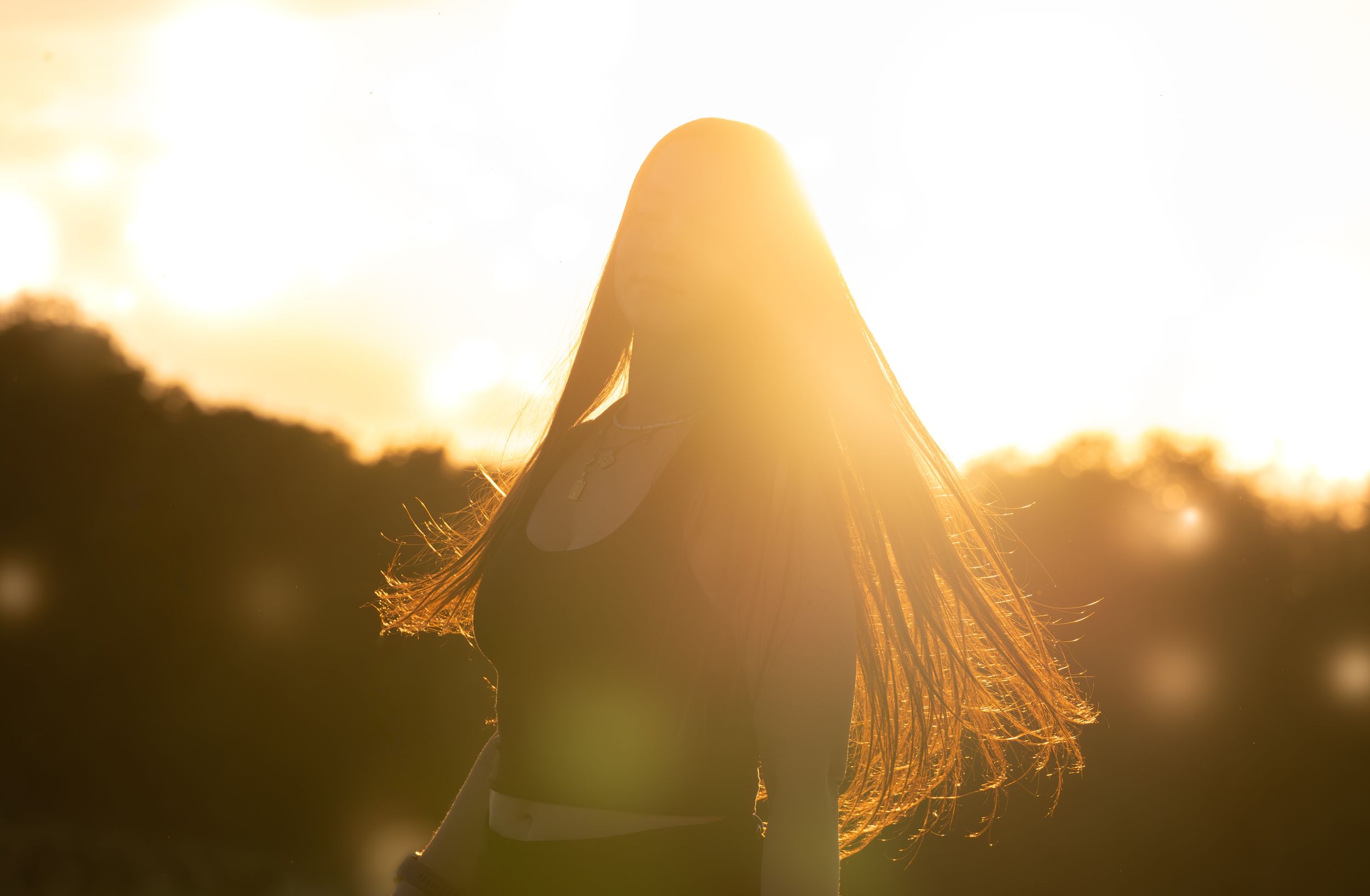 Silhouette of a woman with long hair standing in golden sunset light with glowing backlit hair outdoors.