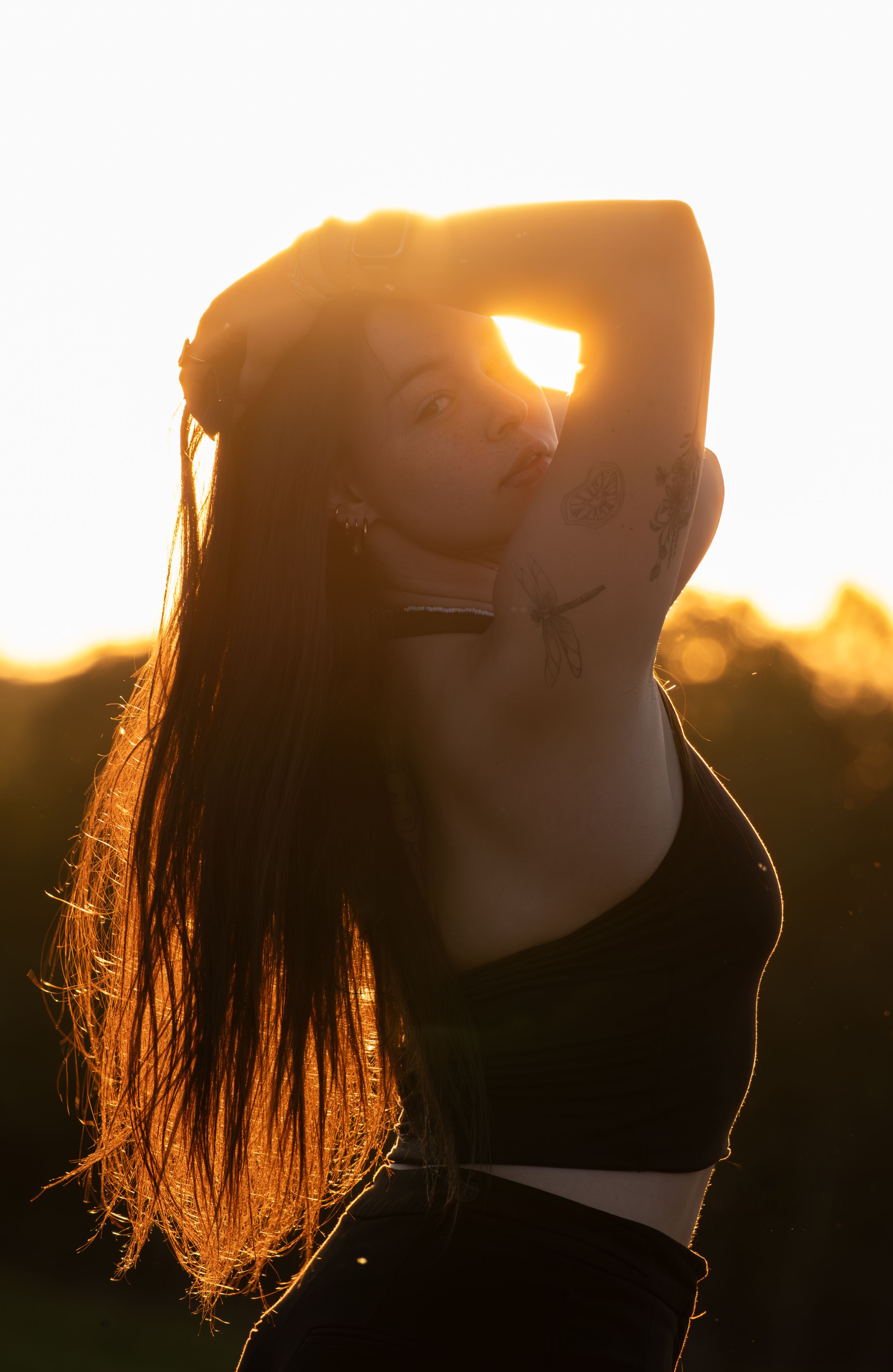 A woman with long hair poses against a stunning golden sunset backlight, wearing a black crop top outdoors.
