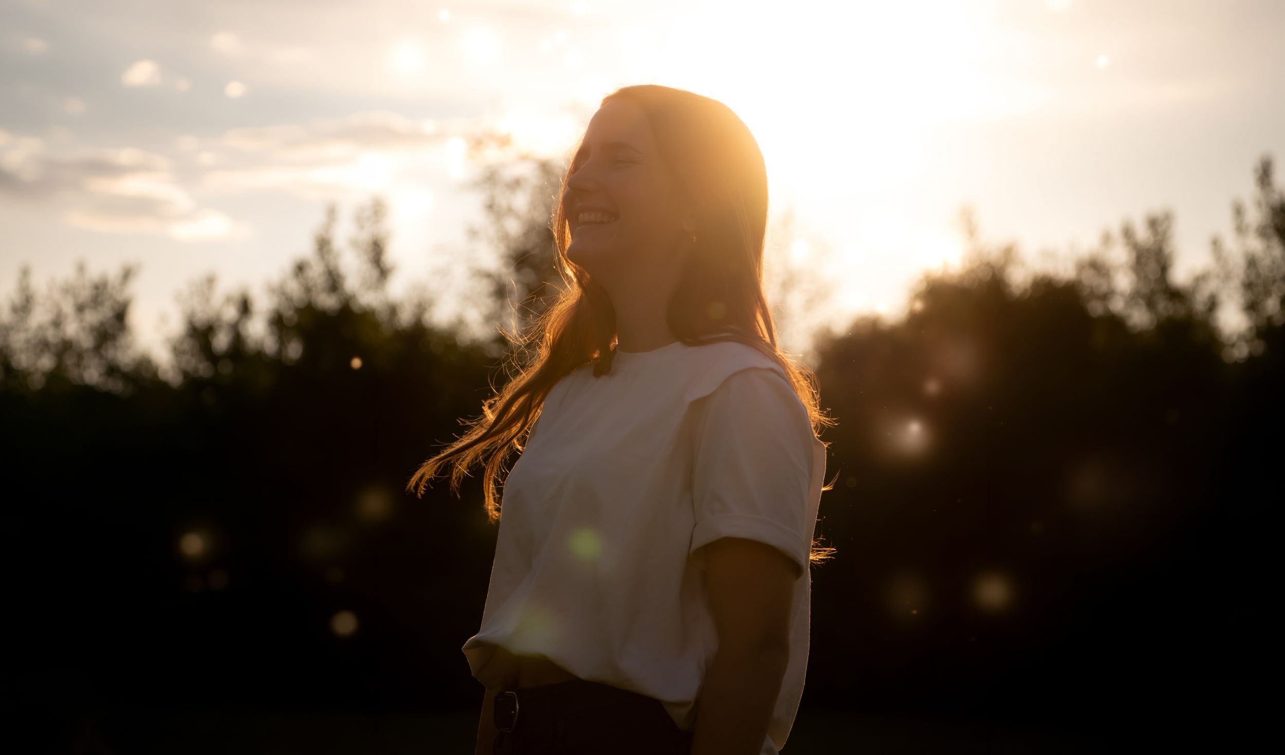 Young woman in white t-shirt basking in golden sunset light outdoors with trees in background.
