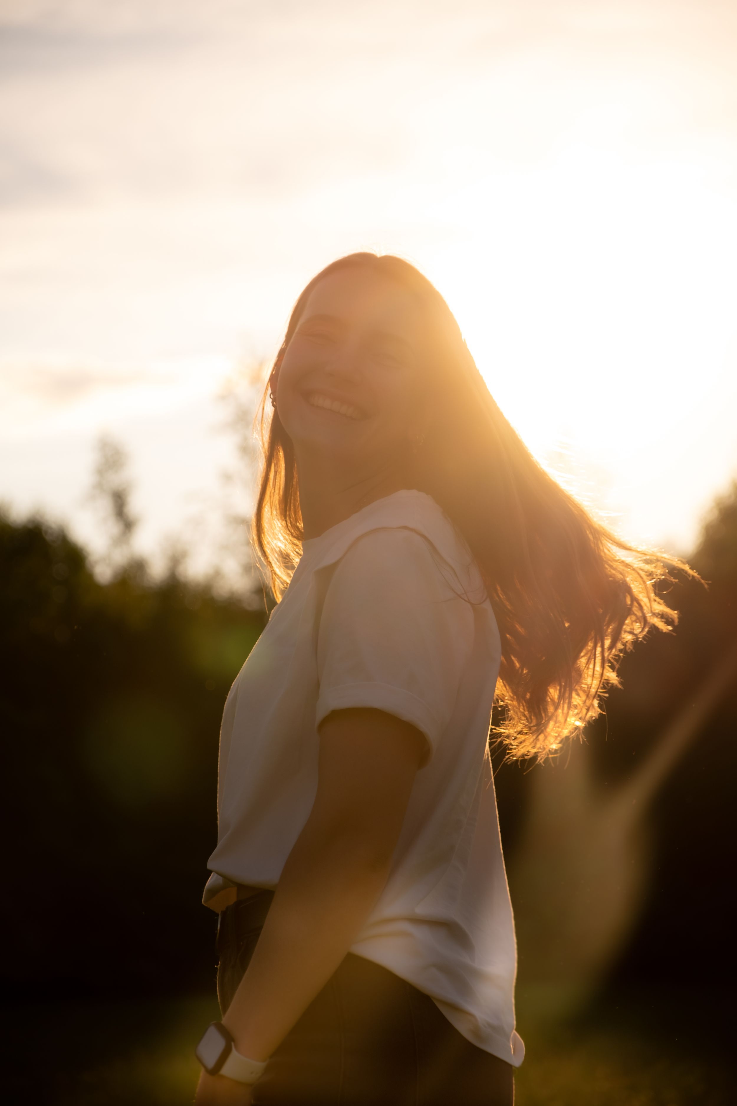Young woman in white t-shirt laughing outdoors during golden sunset with hair blowing in the wind.