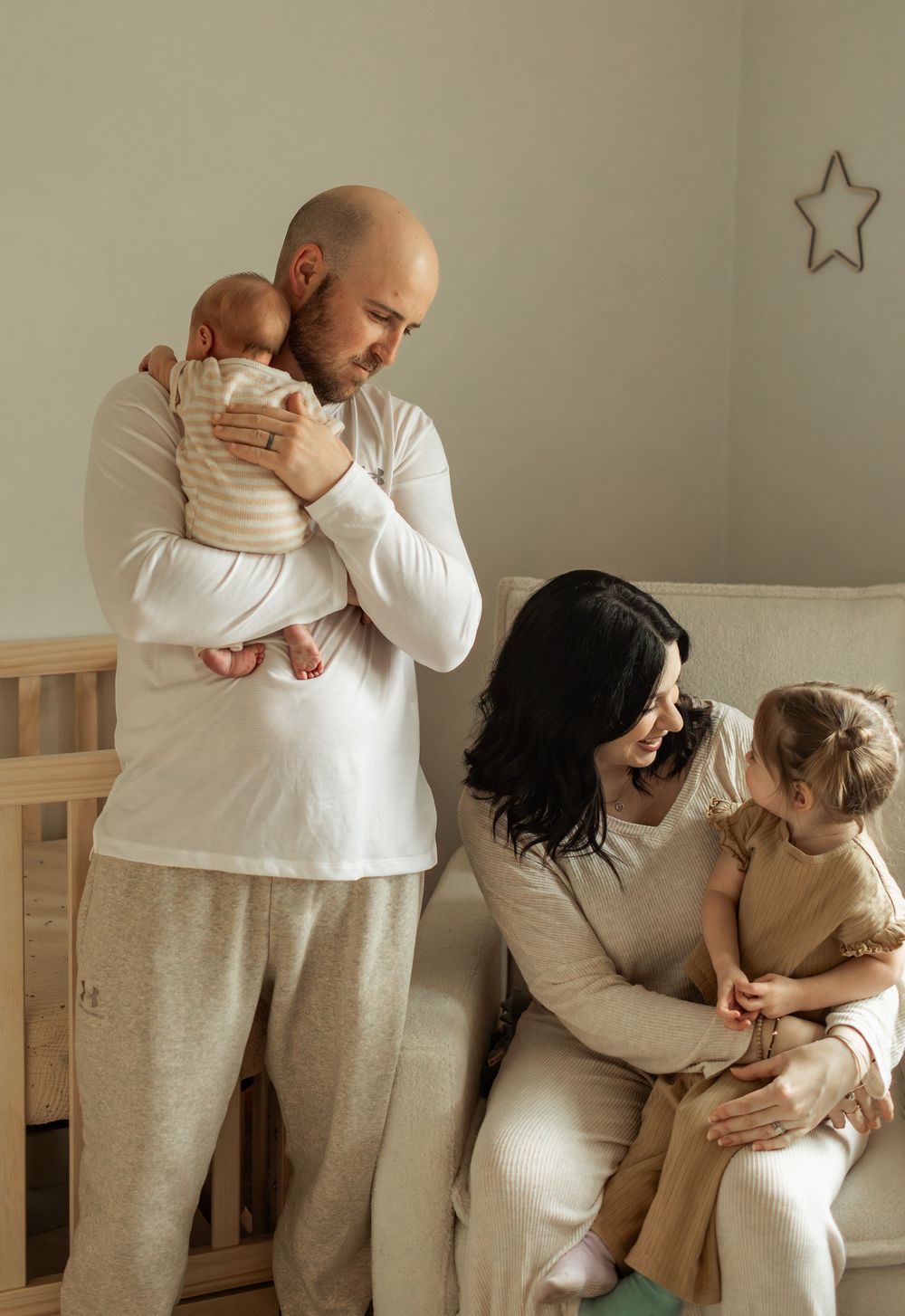 A bald father holds a newborn baby while a dark-haired mother sits with a toddler in a cozy nursery.