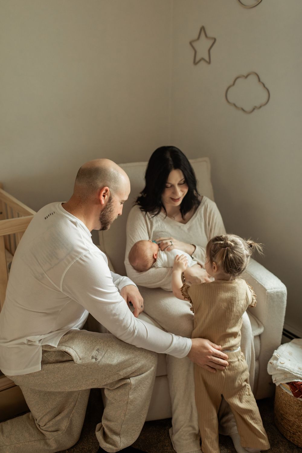 A family of four in neutral tones sits together in a nursery, parents lovingly attending to a newborn and toddler.