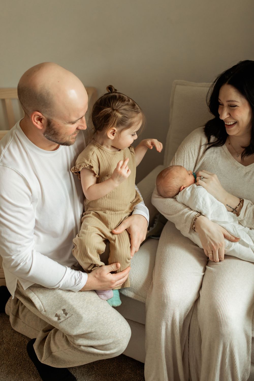 A happy family with a bald father, smiling mother, toddler, and newborn baby sitting together in a cozy room.
