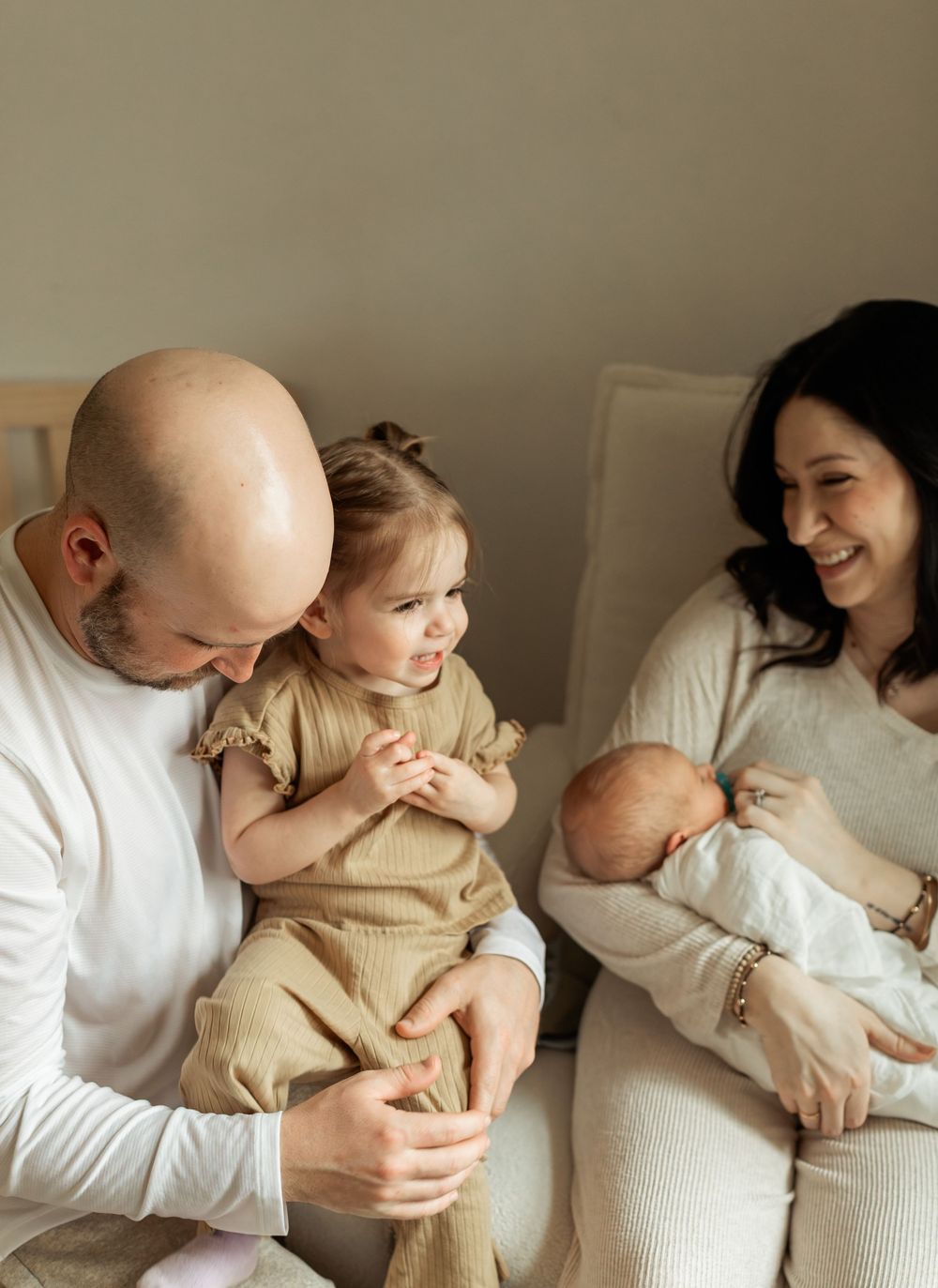 A bald father and dark-haired mother sit together, smiling at their toddler and newborn baby in a warm family portrait.