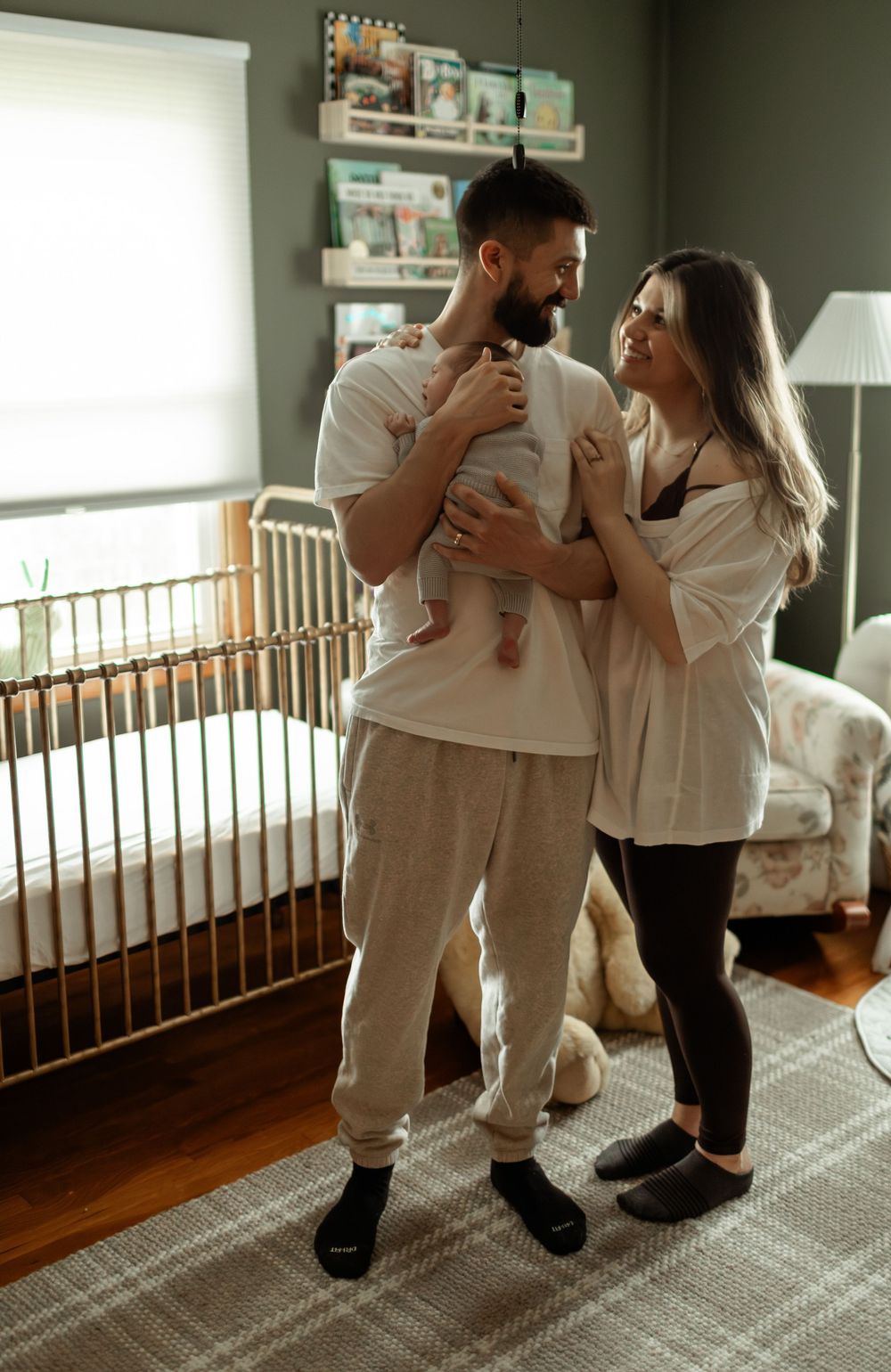Happy couple holding newborn baby in cozy nursery with white crib and warm natural light.