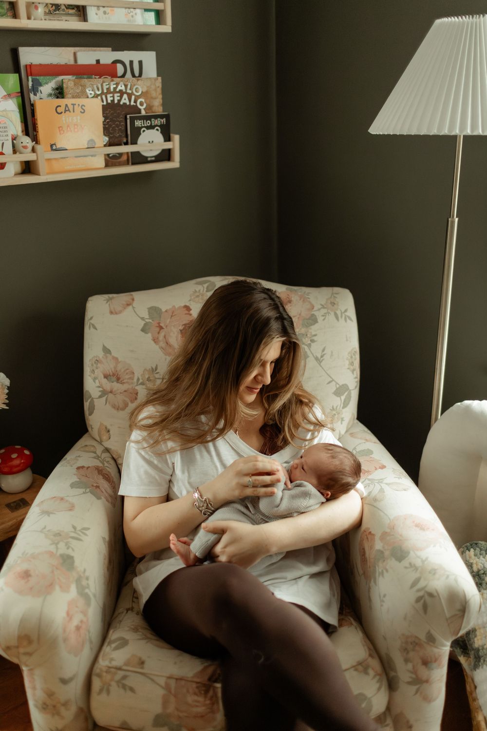 New mother sitting in floral armchair holding newborn baby in cozy nursery room with bookshelf.