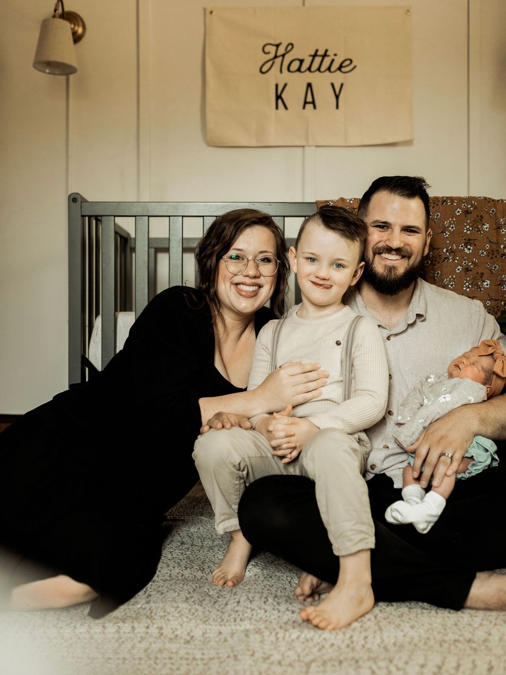 Family of four smiling together in a nursery with a Hattie Kay name banner, including newborn baby and young child.