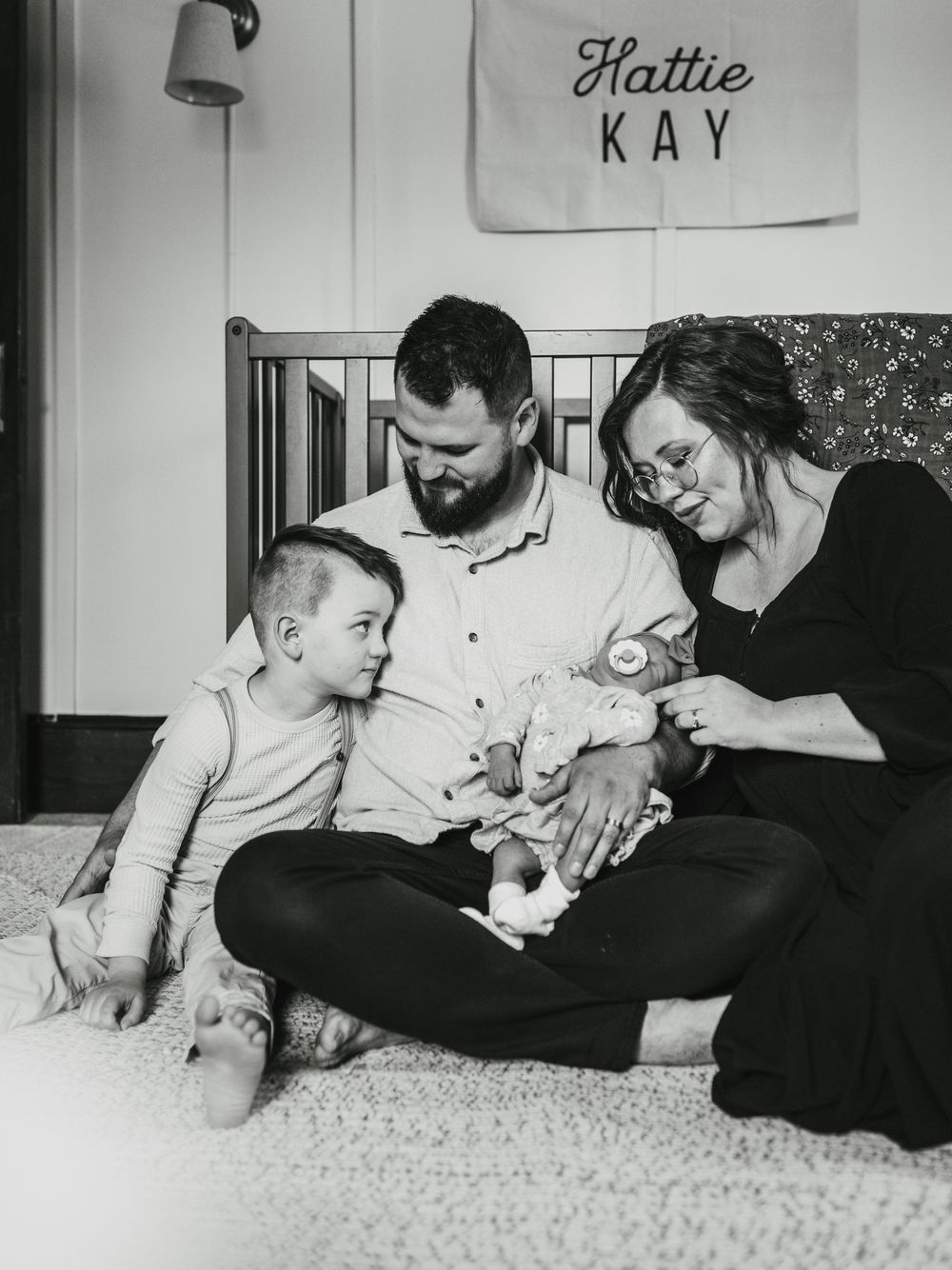 Black and white family photo with parents and toddler admiring newborn baby Hattie Kay in a cozy nursery.