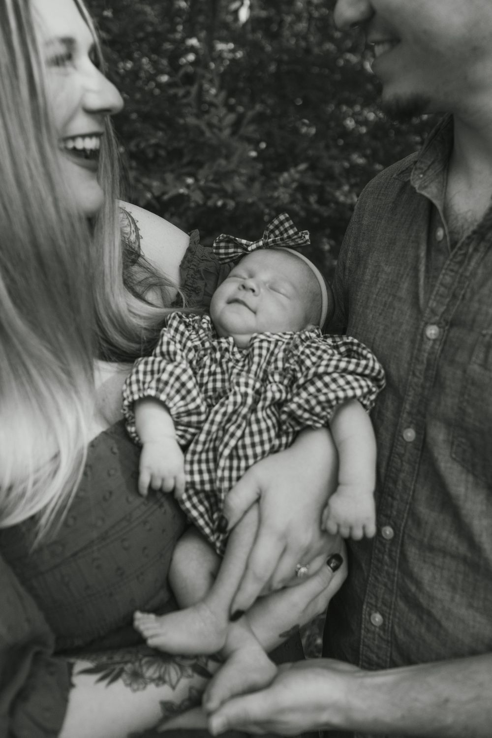 Black and white photo of smiling parents holding their newborn baby dressed in plaid outfit with bow headband outdoors.