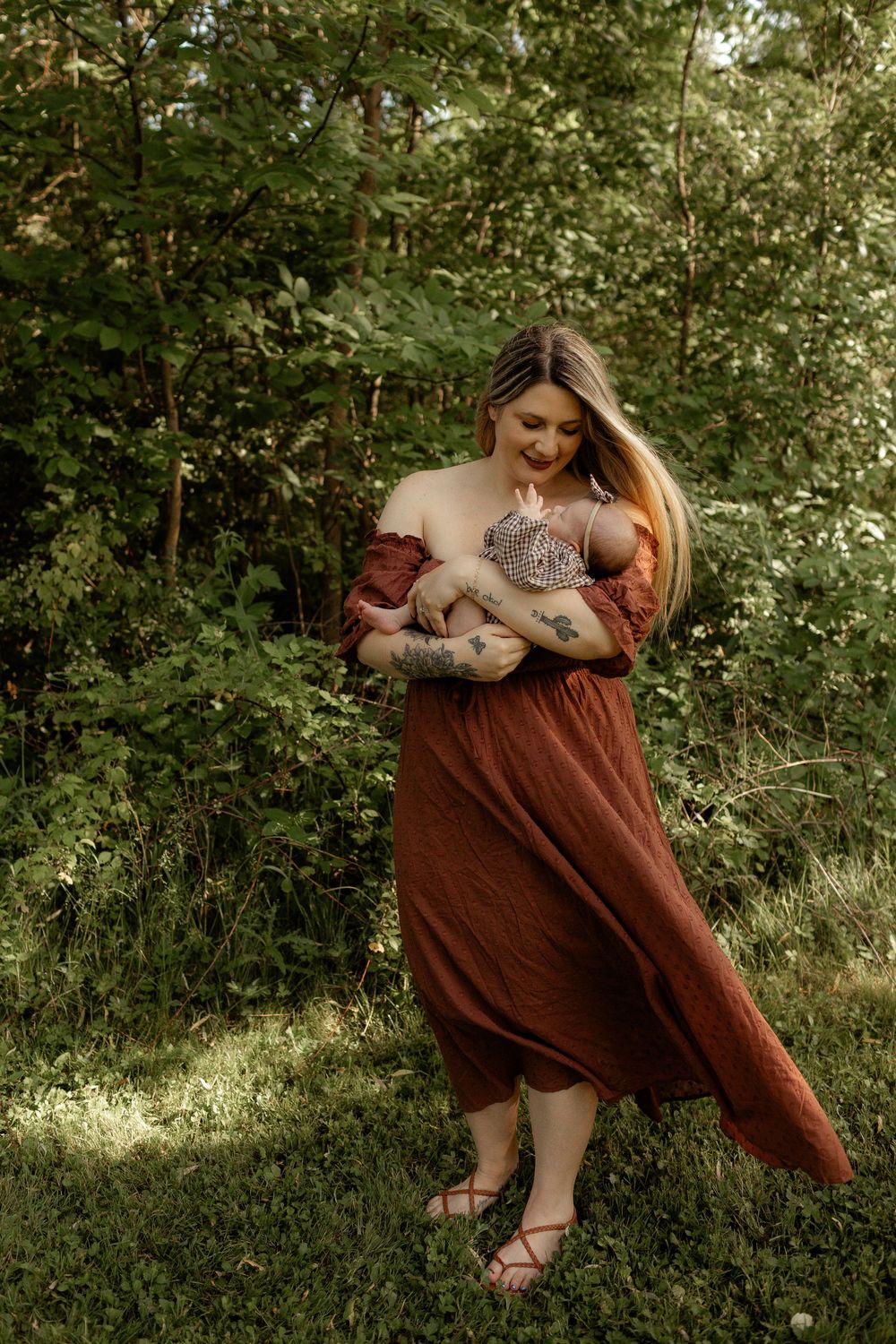 A tattooed woman in a rust-colored dress holds her newborn baby outdoors in a lush green forest setting.