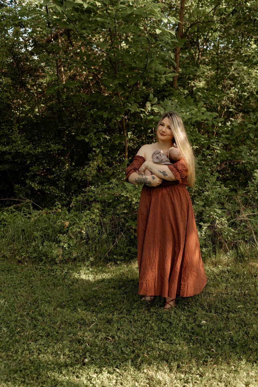 Woman in rust-colored maxi dress holding newborn baby outdoors in lush green forest setting.