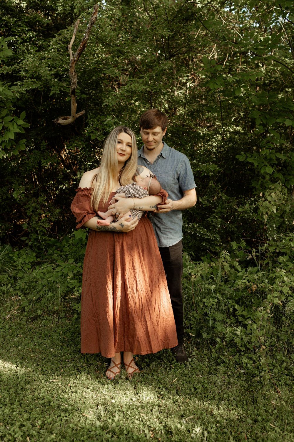 Young couple in lush garden holding newborn baby, woman in rust dress, man in blue shirt, outdoor family portrait.