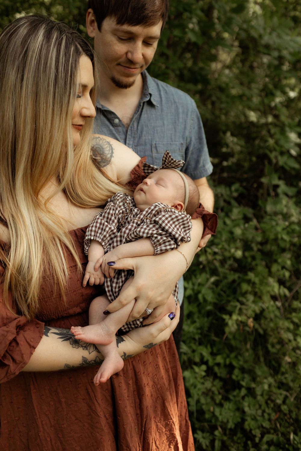 New parents lovingly cradle their sleeping newborn baby outdoors, mother in rust dress with tattoos, father in blue shirt.