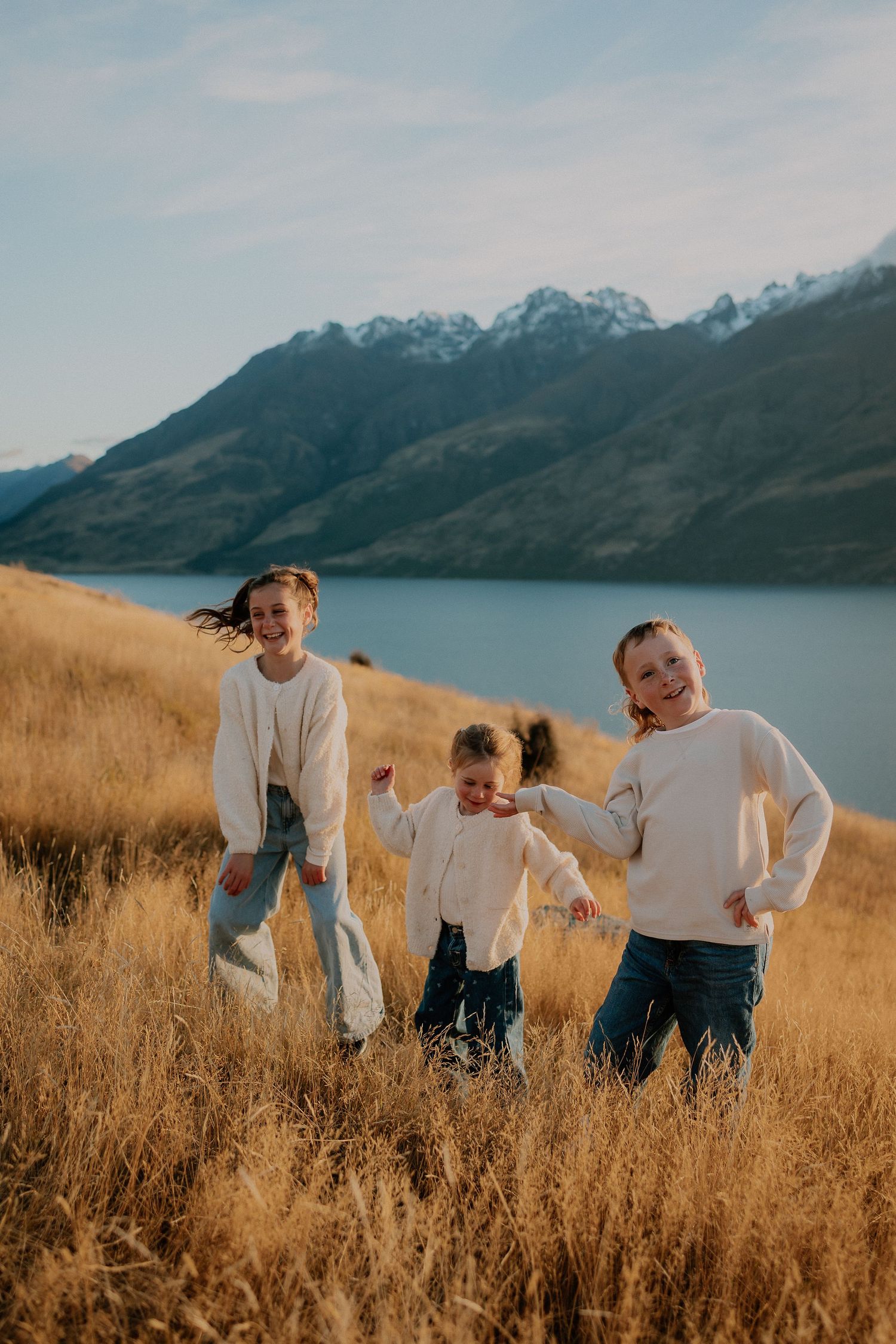 Three children laughing and playing in golden grass fields with a scenic lake and snow-capped mountains backdrop.