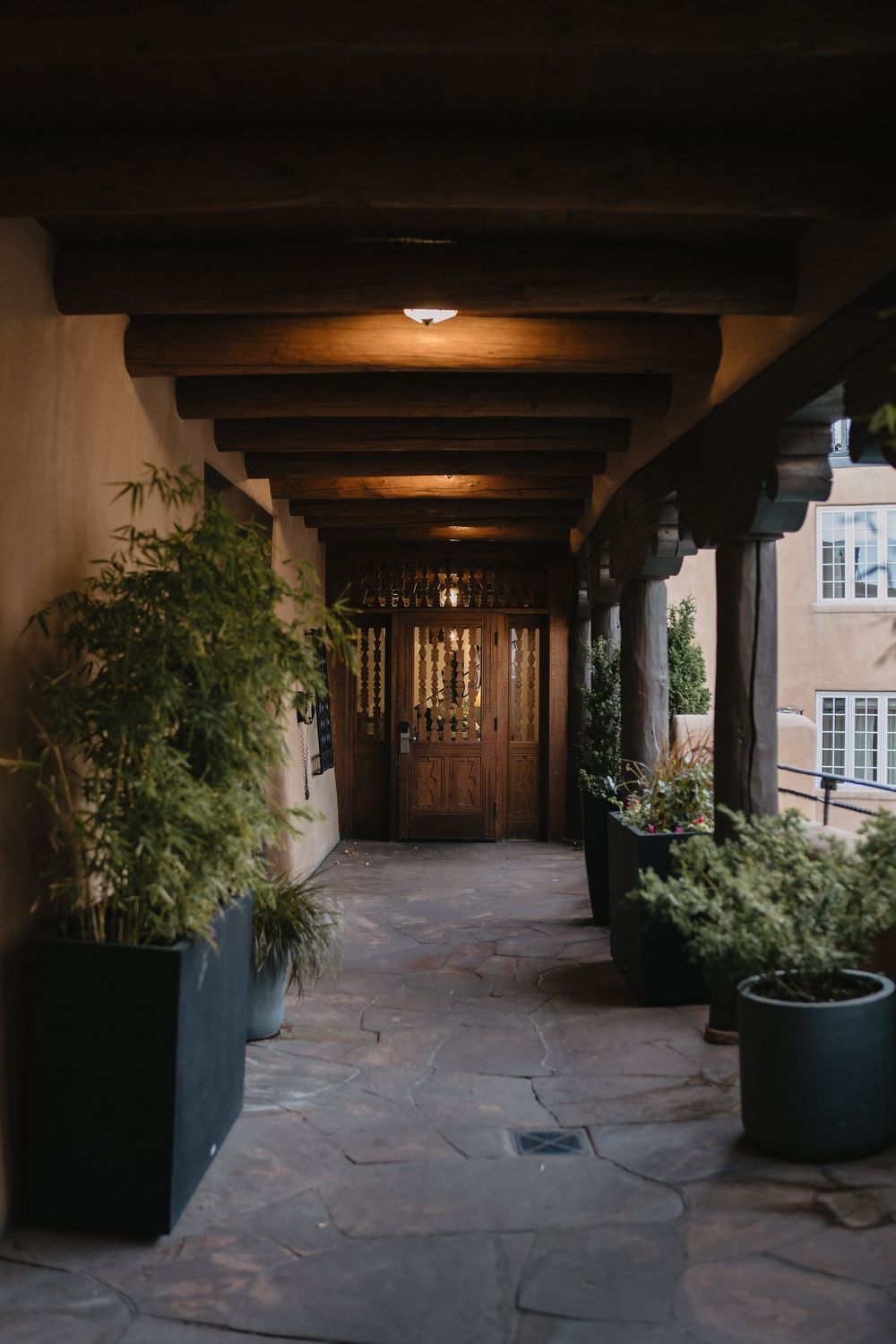 A dimly lit Southwestern-style covered walkway with wooden beam ceiling, stone floor, potted plants, and ornate wooden door.
