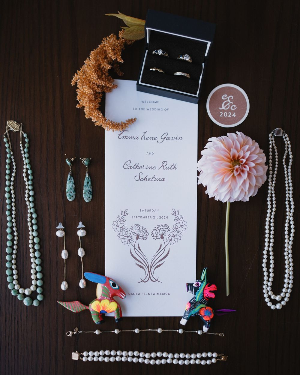 Wedding flat lay with invitation, rings, pearl necklaces, turquoise jewelry, pink dahlia, and colorful folk art figurines.