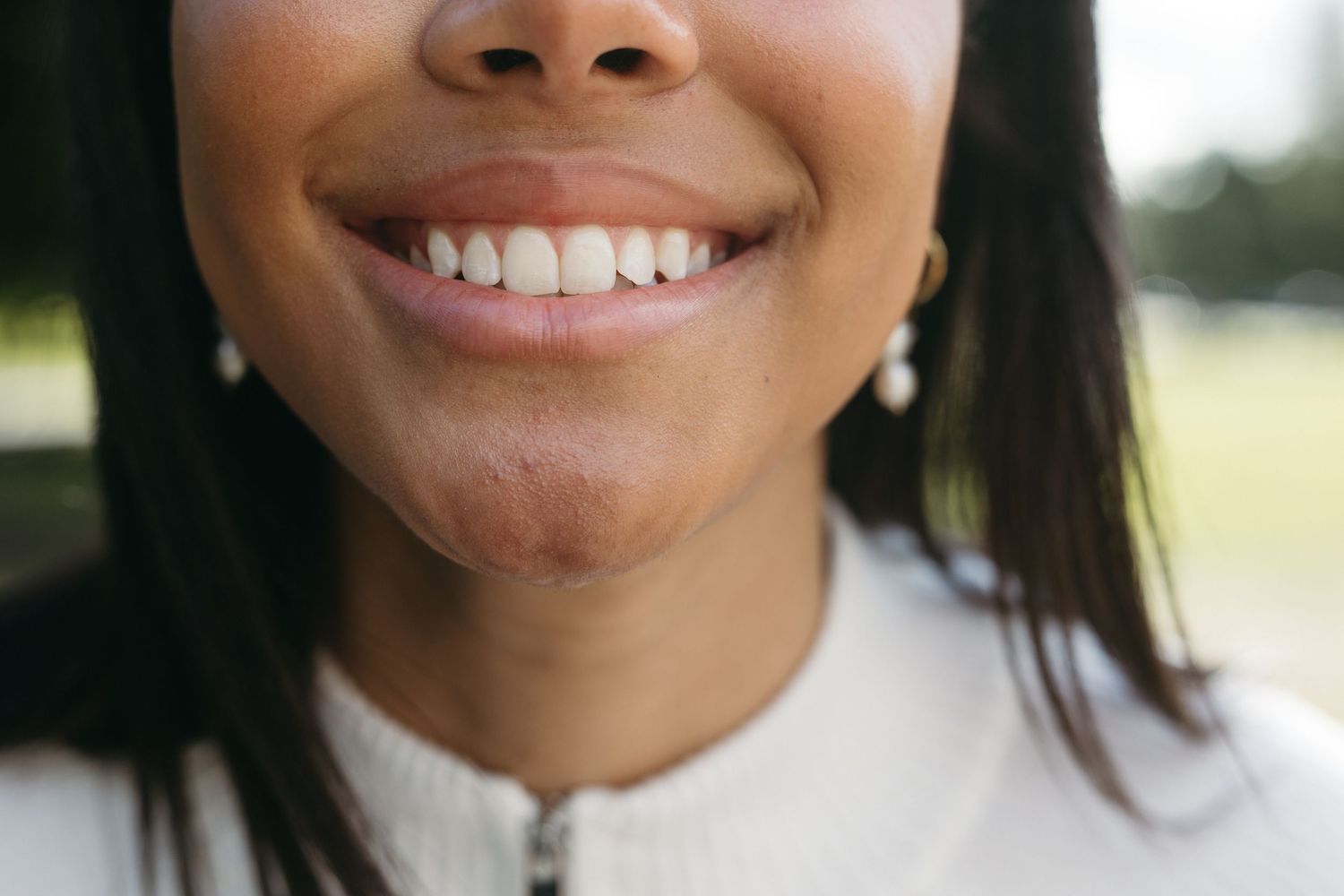Close-up of a smiling young woman with white teeth, dark hair, and pearl earrings wearing a white top outdoors.