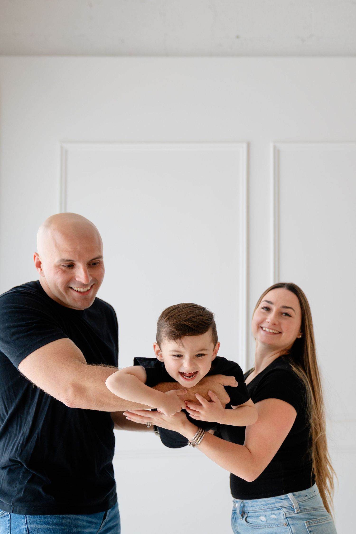 Happy family with bald father, mother, and young son in black outfits playing together against white wall.