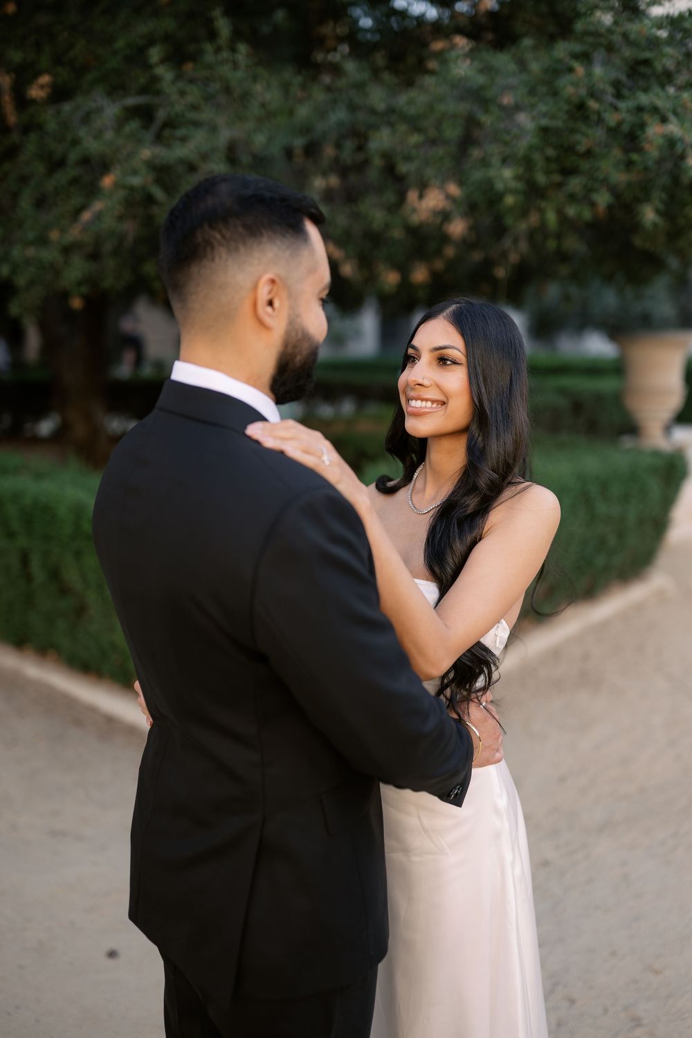 A smiling bride in a white gown embraces her groom in a black suit outdoors in a lush garden setting.