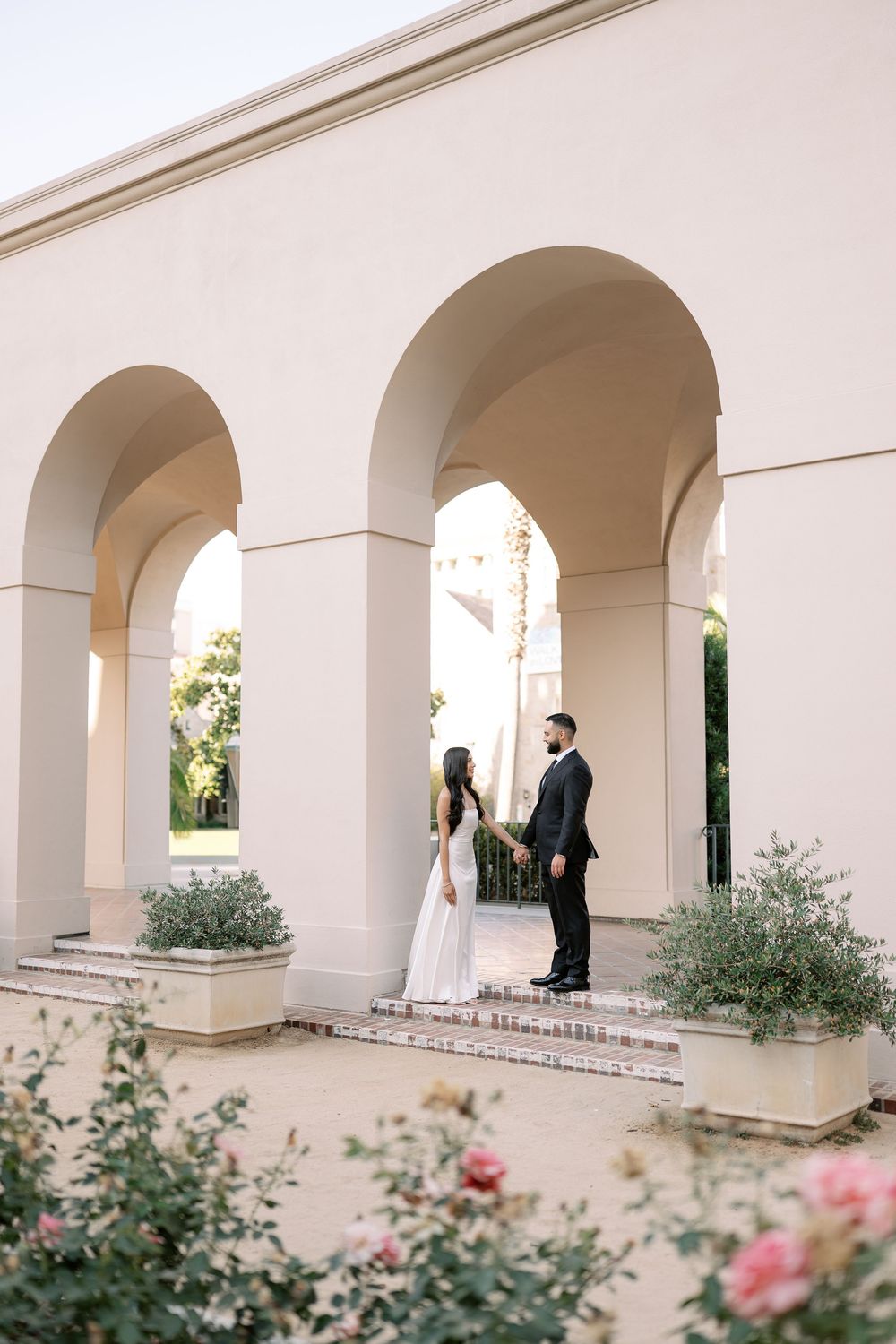 Bride and groom hold hands under elegant arched colonnade with rose gardens at a wedding venue.