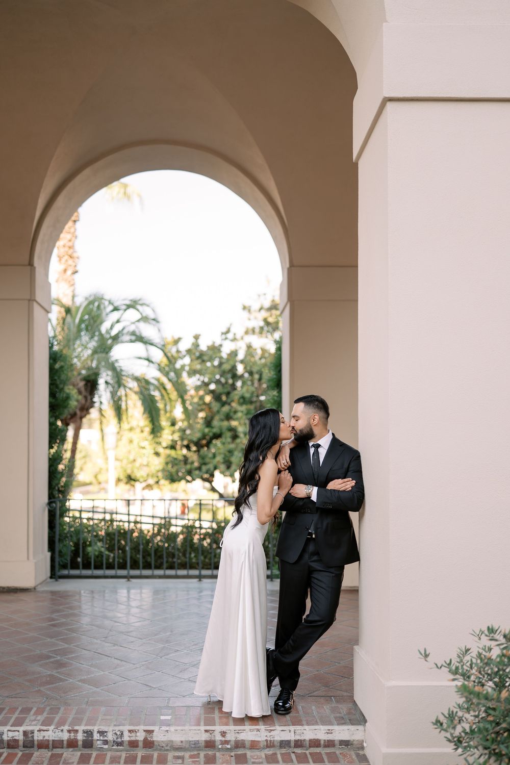 Elegant couple in wedding attire sharing a romantic moment under an arched colonnade with palm trees in the background.