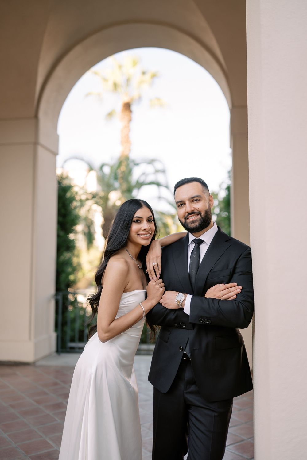A couple poses elegantly under an arch, bride in white gown and groom in black suit, with palm trees in background.