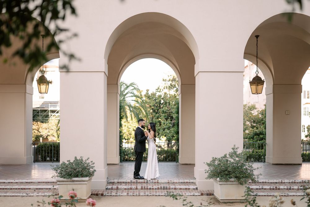 Bride and groom share a romantic moment under elegant white arches surrounded by lush greenery at an outdoor wedding venue.