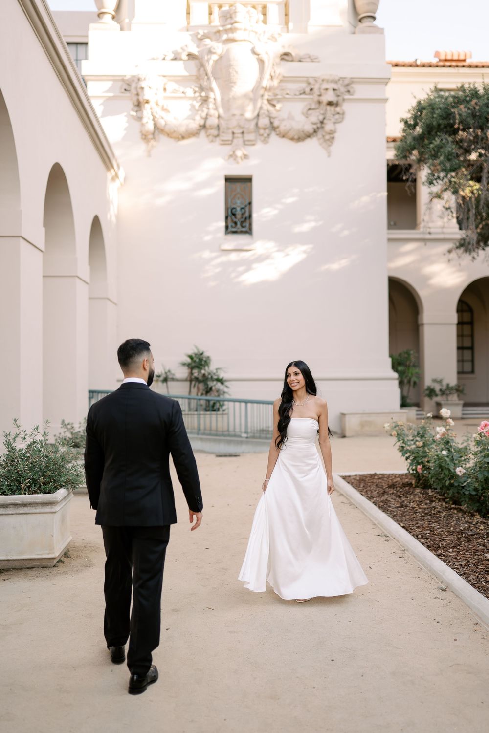 Bride in white strapless gown and groom in black tuxedo walking through elegant courtyard of white building.