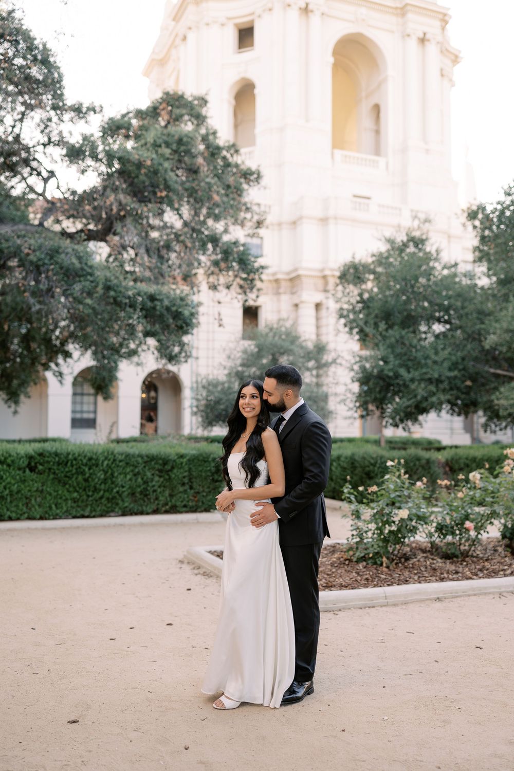Elegant couple in wedding attire posing in front of a grand building with lush greenery and manicured hedges.