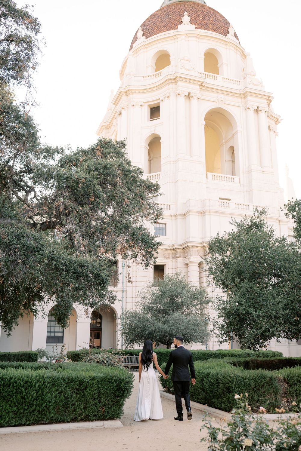 Wedding couple walks hand in hand toward ornate Pasadena City Hall tower surrounded by lush hedges.