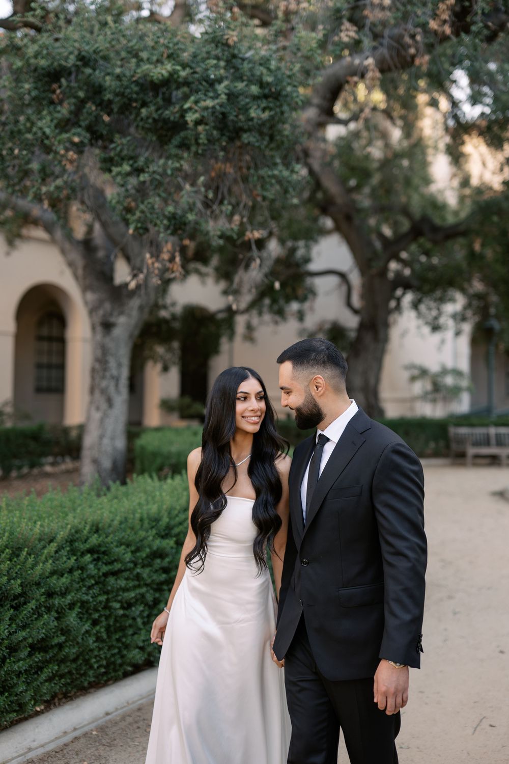 Elegant couple in formal attire walking through a garden courtyard, woman in white gown and man in black suit.