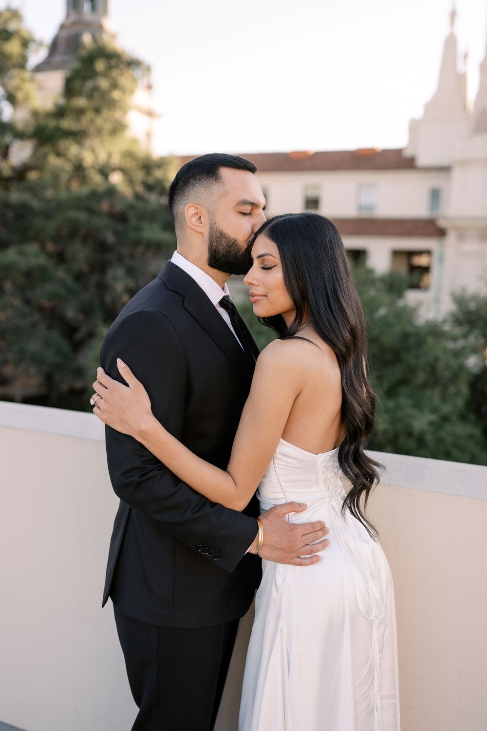 Romantic couple embracing on a rooftop terrace, groom in black suit kissing bride in white dress.