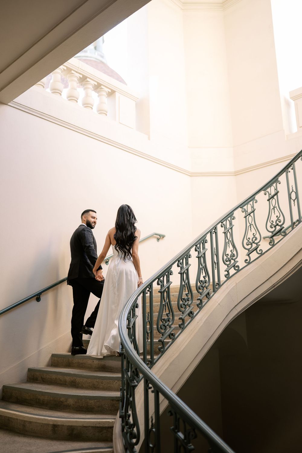 Elegant couple holding hands ascending ornate spiral staircase in white gown and black suit.