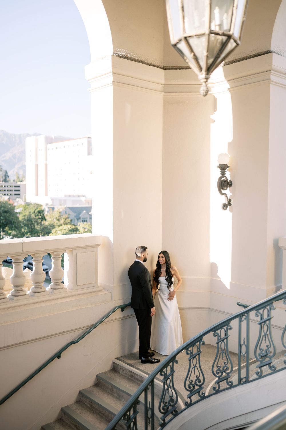 Elegant bride and groom share romantic moment on grand staircase of luxury wedding venue balcony.